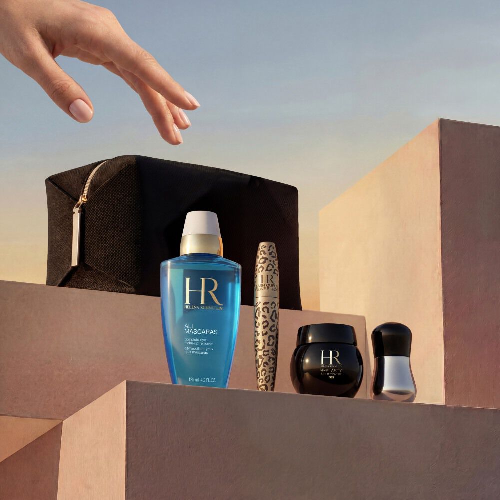 Une main elegante s'approche d'une selection de produits Helena Rubinstein disposes sur des blocs en pierre beige sable minimalistes. L'ensemble est presente sur fond de ciel dore au coucher du soleil et comprend une pochette de voyage noire, le demaquillant yeux bleu 'All Mascaras', un mascara Lash Queen Feline Blacks a motif leopard, un petit pot de creme Re-Plasty noir et un outil applicateur argente.​
