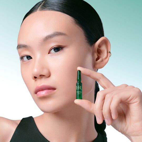 Gros plan sur le visage d'une femme tenant delicatement entre ses doigts une ampoule verte Powercell [EXO] Skinmunity Helena Rubinstein, sur un fond degrade vert et blanc.​