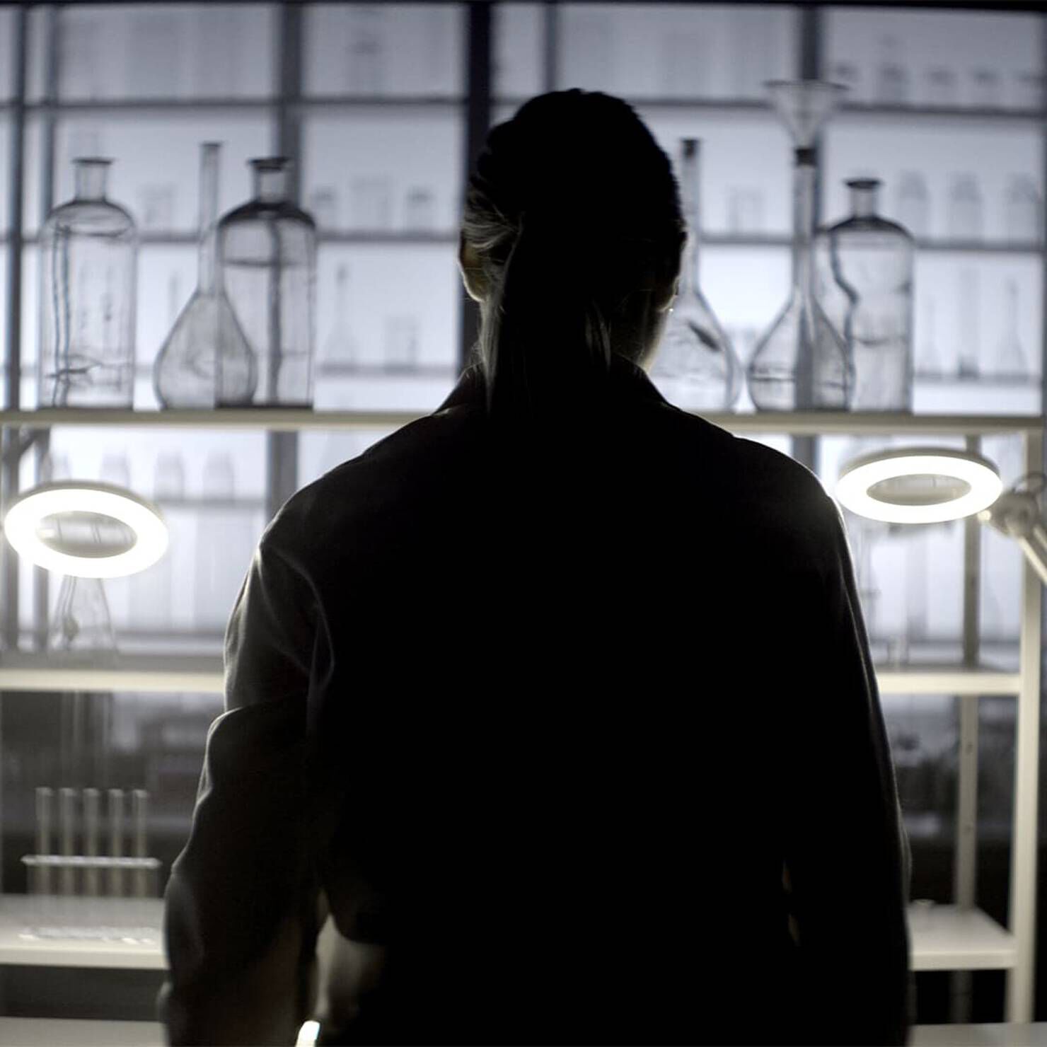 Une silhouette de femme se tenant devant des instruments de laboratoire, eclairee par des anneaux lumineux.