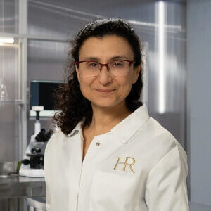 Scientifique en blouse blanche avec des lunettes, souriant, se tenant devant un microscope en laboratoire moderne.
