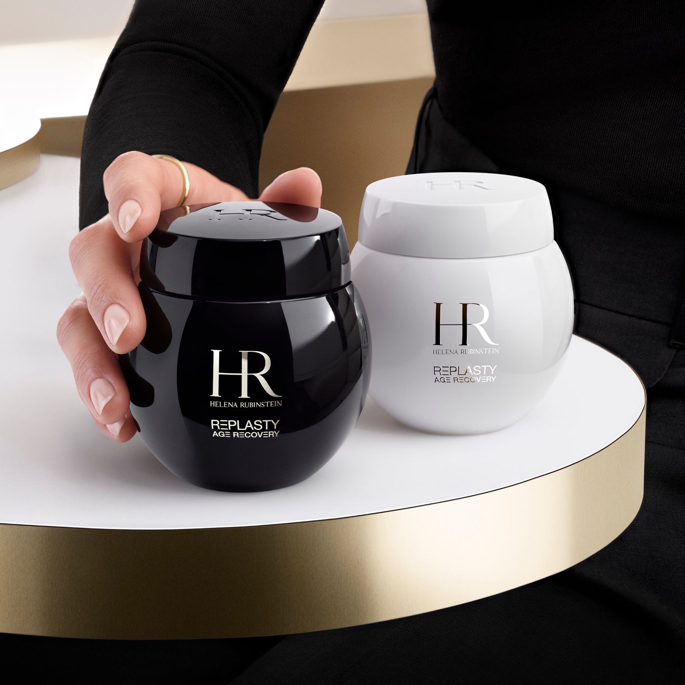 Une main saisit un pot noir de creme Helena Rubinstein Re-Plasty Age Recovery, un pot blanc assorti a ses cotes, sur une plateforme doree.