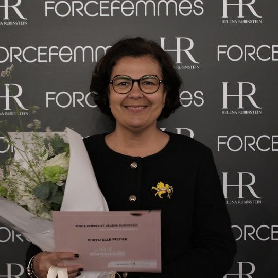 Une femme souriante, aux cheveux courts et fonces, porte des lunettes et une veste violette, tenant un bouquet de fleurs blanches devant un mur de logos.