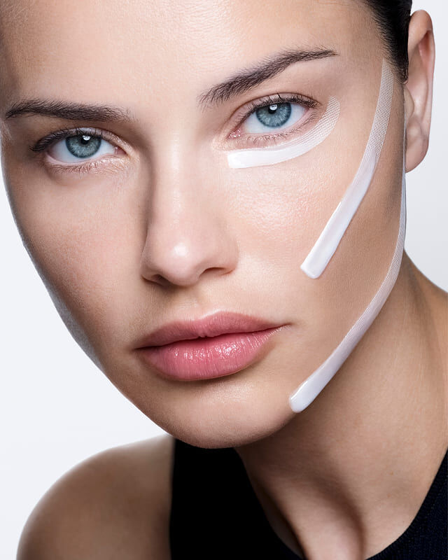 Adriana Lima avec de la creme blanche et des bandes appliquees sur son visage, montrant un effet lifting visible.