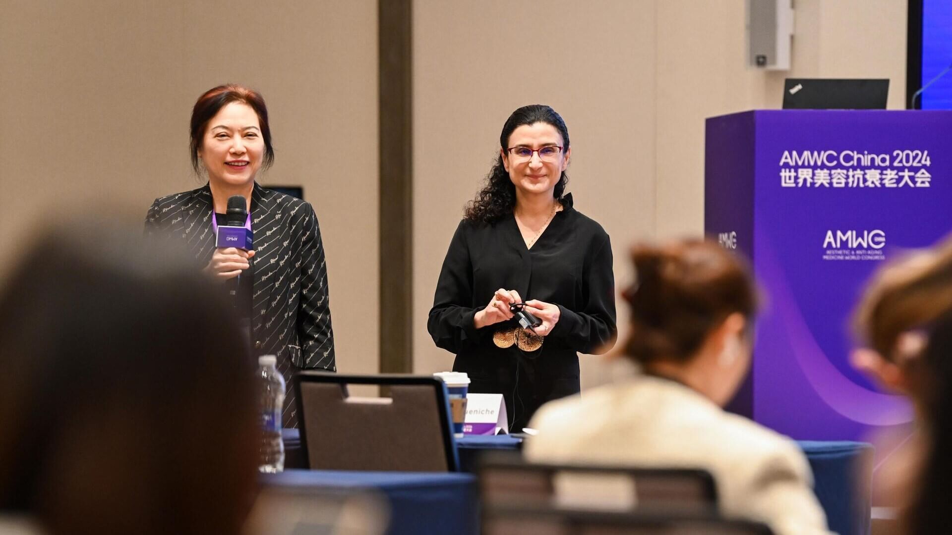 Deux femmes souriantes se tiennent devant un public, lors de l'evenement AMWC China 2024, avec un podium en arriere-plan.