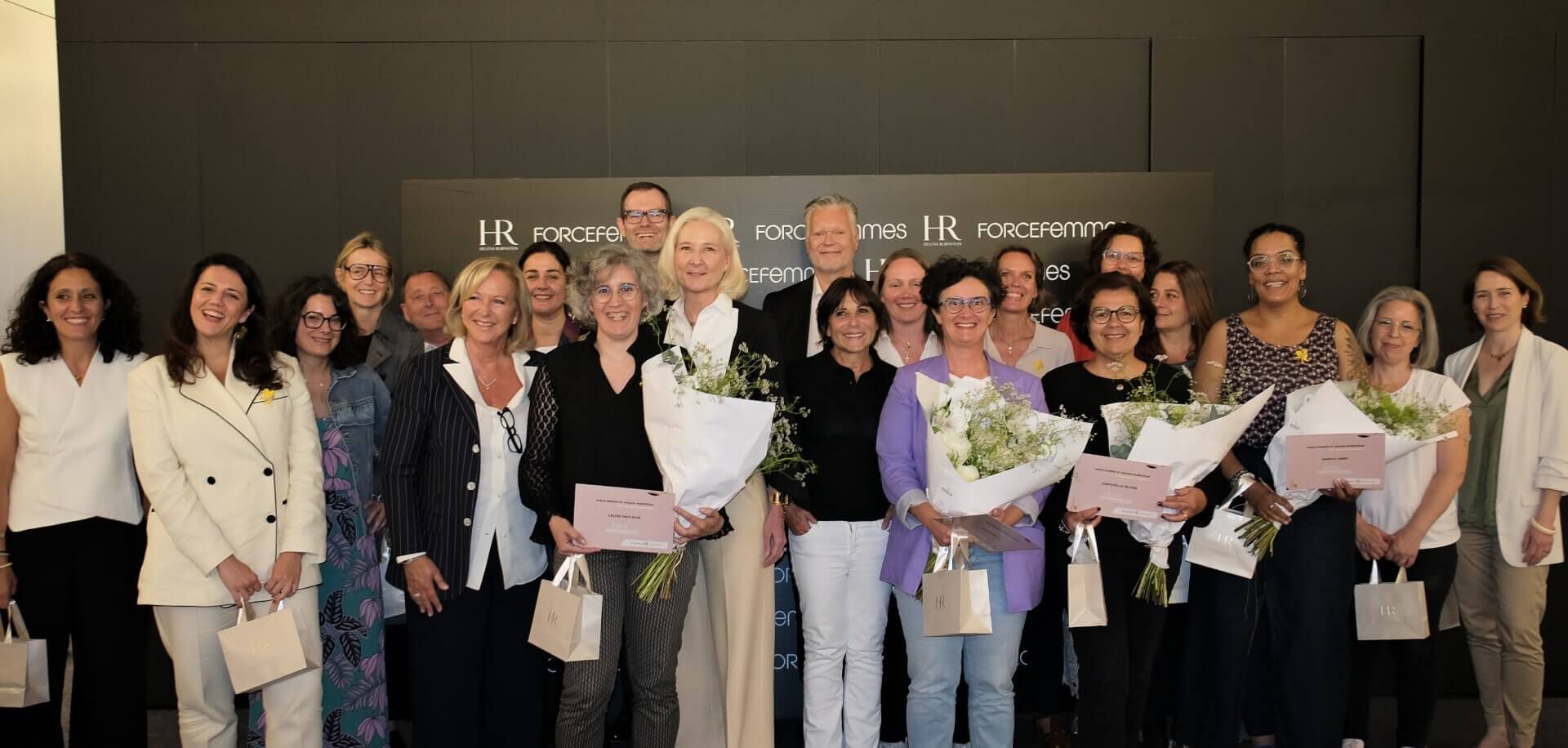 Grand groupe de personnes, majoritairement des femmes, certaines tenant des bouquets et des sacs, posant joyeusement devant un mur noir avec les logos "HR" et "FORCEfemmes".