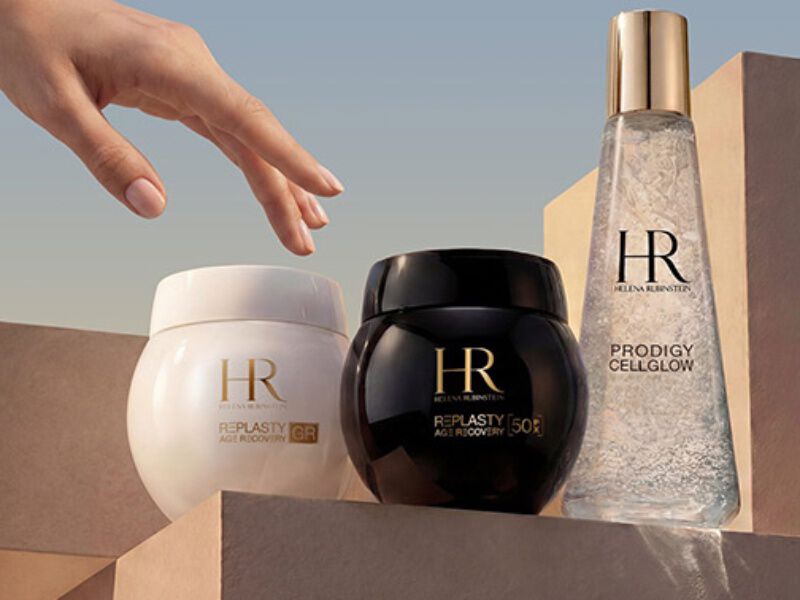 Une main se tend vers trois produits Helena Rubinstein, mis en valeur par un design elegant et un eclairage tamise.