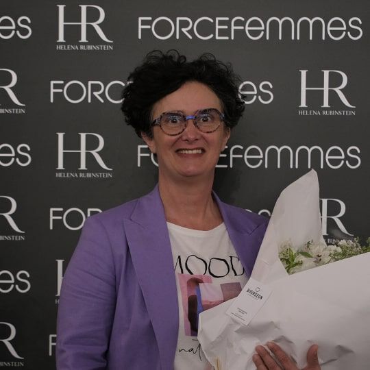 Une femme souriante, aux cheveux courts et fonces, porte des lunettes et une veste violette, tenant un bouquet de fleurs blanches devant un mur de logos.
