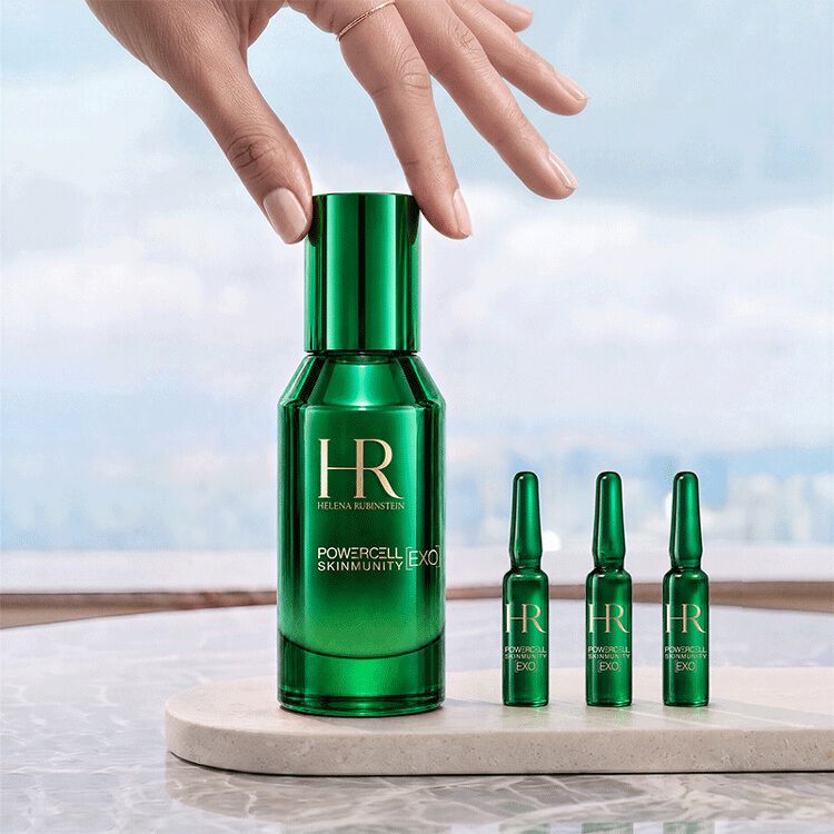Une main touchant le flacon vert du serum Powercell [EXO] Skinmunity Helena Rubinstein, pose a cote de trois ampoules [EXO] Shots sur un support en marbre devant un paysage lumineux. ​