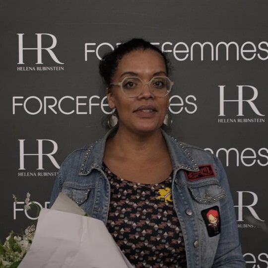 Une femme souriante, aux cheveux fonces attaches, porte des lunettes rondes et une veste en jean, tenant un bouquet de fleurs blanches devant un mur de logos.