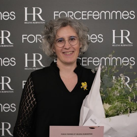 Une femme aux cheveux gris boucles et lunettes sourit en tenant un bouquet de fleurs. Elle porte un haut noir avec un detail en dentelle. Fond de logos.