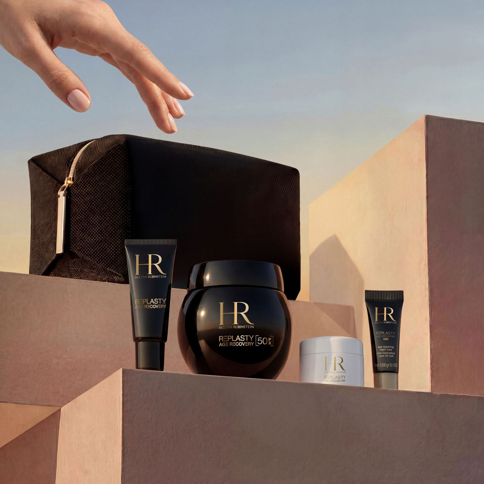 Une main elegante s'approche d'un coffret cadeau Helena Rubinstein, dispose sur des blocs de pierre sablonneux beige sur fond de ciel de coucher de soleil, comprenant une pochette de voyage edition limitee noire et la gamme Replasty Age Recovery : Eye Repairing Night Care, Day Cream, Night 50PX et Body Repairing Care.​